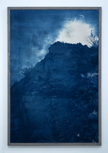 The images is vertical and  a cyanotype. It's of a mountain or rocky hill. The sun is setting behind it and rays are coming from the top. There are hints of rocks and plant life.  