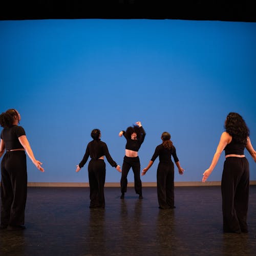 A group of five female dancers performing a Senior Dance Capstone Project. The central dancer who is Iranian and Mexican, stands with arms raised in fists towards the audience, surrounded by four dancers of Black, Latinx, and Asian decent, forming a V shape. All dancers are dressed in black attire against a blue backdrop.