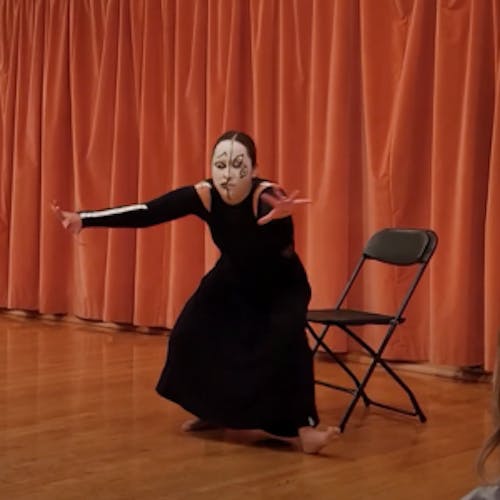 A dancer sits on a chair in the middle of the room, giving her back to the audience. She is wearing a black skirt and a black shirt with long sleeves. Her limbs and spine are outlined with white tape, making them look like skeletons. The music has an eerie tone, the sound of an opening and closing door repeats repeatedly. The extends her arms with difficulty to the sound of the door. She turns around, hiding her face. Voices in a choir make dissonant harmonies. The sound of tapping on a guitar comes and goes in sporadic flourishes. Her face is painted white. One side of her face has her mouth screaming, her eyes wide open, and an exaggerated brow giving it a frown. The other side of her face has two big eyes drawn with black and tilted diagonally, giving it a surrealistic feel. She continues to make fast and precise arm movements to the tapping rhythm. Her torso undulates in pain, while her feet don't seem to find her footing. Her arms create lines, circles, and accents to the music. Finally, the sound of rain comes as she settles back into the characters backing up until the music is gone. 