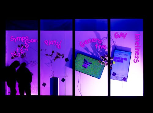 The photograph captures a nighttime art exhibition presented in a display window. Two young women, likely UCLA students, stand in the lower left corner, gazing down as if reading. One of them is wearing a hoodie. The display window is divided into four sections.  In the first section, the words "Sympozoom" and "Natural" are written in large, bold pink letters using the Comic Sans font on the glass. The arrangement of the letters in "Natural" is jumbled. There are additional smaller vinyl texts, but they are too tiny to decipher. Behind the glass, a dried branch with leaves can be seen.  The second section features the word "Playful" on the glass in the same font and size as the previous words. Another dried branch with leaves is visible behind the glass. Below the branch, a small sculpture rests on a slim pedestal, consisting of an unusually-shaped tree limb adorned with two 3D-printed plastic kinetic robots. The robots' arms extend outward, attached to the branch's twigs.  In the third section, the prominent word on the glass is "Generative." A large TV screen is visible through the glass, displaying a floral-like 3D figure with human eyes against a predominantly green backdrop speckled with multicolored dots.  The fourth section's glass features the words "GAY" and "loneliness." A sizeable, slanted, vertically-mounted TV can be seen through the glass, showing a web user interface. However, the text on the screen is too minuscule to read.  Additionally, six paper-printed QR codes are affixed to the glass panels, distributed across all four sections. The overall color scheme of the image is tinged with purple and blue hues.