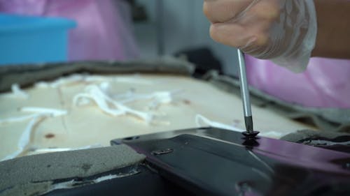 A still from a video showing a close up of a pair of gloved hands holding a screwdriver and disassembling an office chair. 