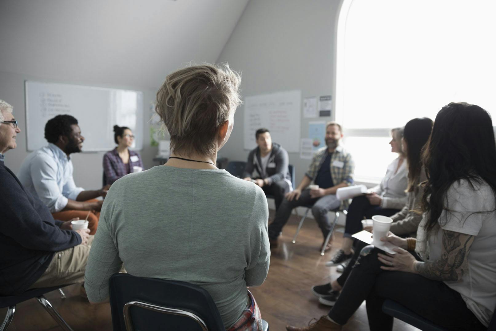 A support group. 9 adults are sitting in a circle in a classroom. There are two whiteboards in the background and a large window to the right. Each person in the circle has a name tag on. They are all holding either a cup or a clipboard or both. Most people are smiling.