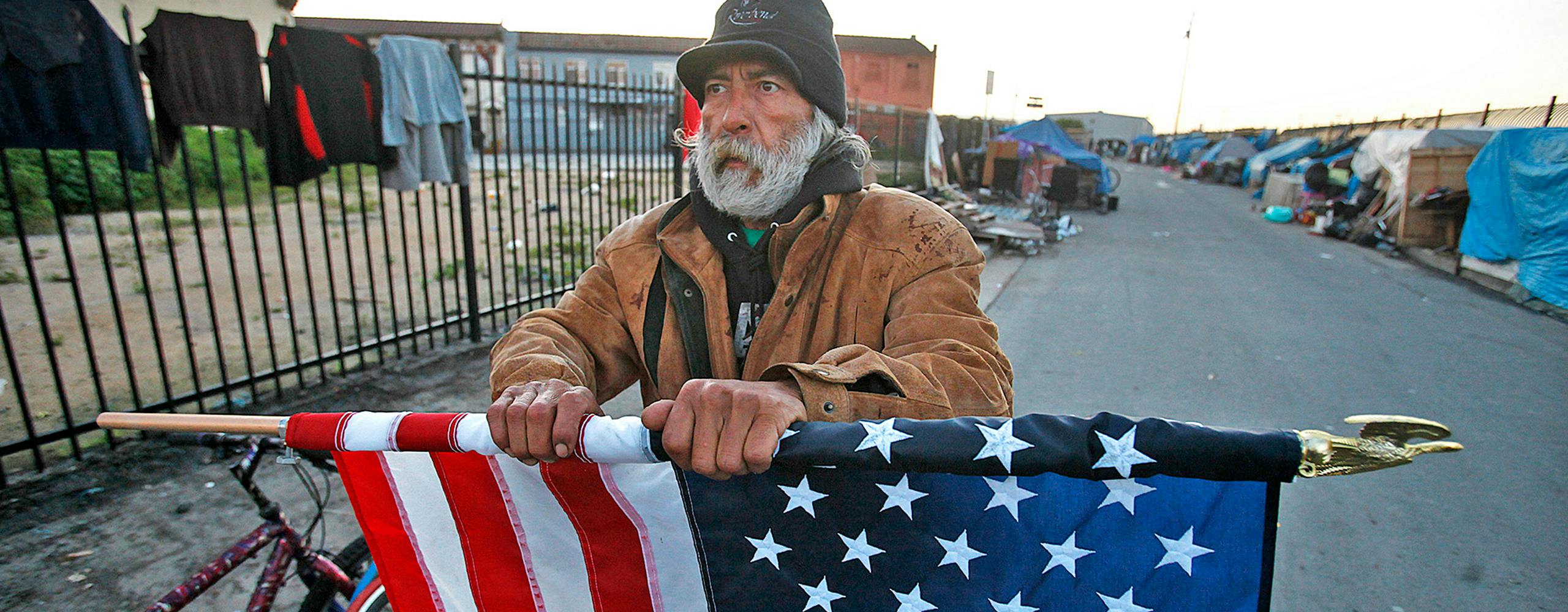 A homeless person holds a slightly rolled American flag. They appear to be standing in Skid Row Los Angeles, a street with many homeless tents lining the street. There is a fence with clothes hanging on it to the left of the homeless man, as well as two bikes to his direct left.