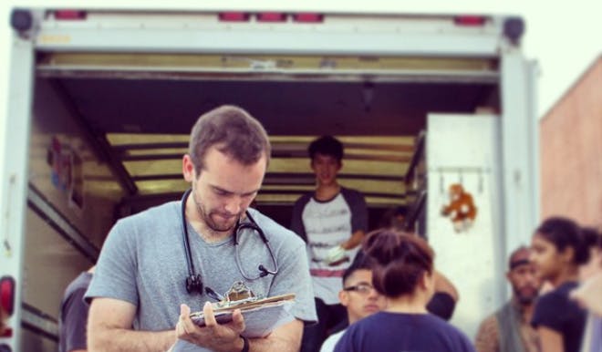 A man with a stethoscope around his neck is looking at a clipboard. He is holding a pen in his hand. Behind him is the back of an ambulance with one kid standing and smiling inside, and four children standing in front of the ambulance behind the man.