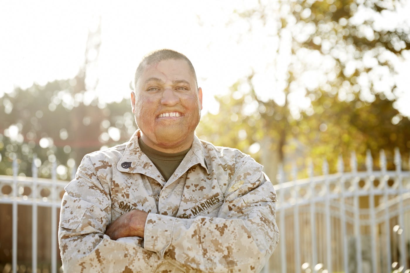 A Marine veteran stands with their arms folded across their chest, wearing a camo jacket. The words SAN and MARINES are visible on the jacket. In the background there is a fence with blurry trees behind it.