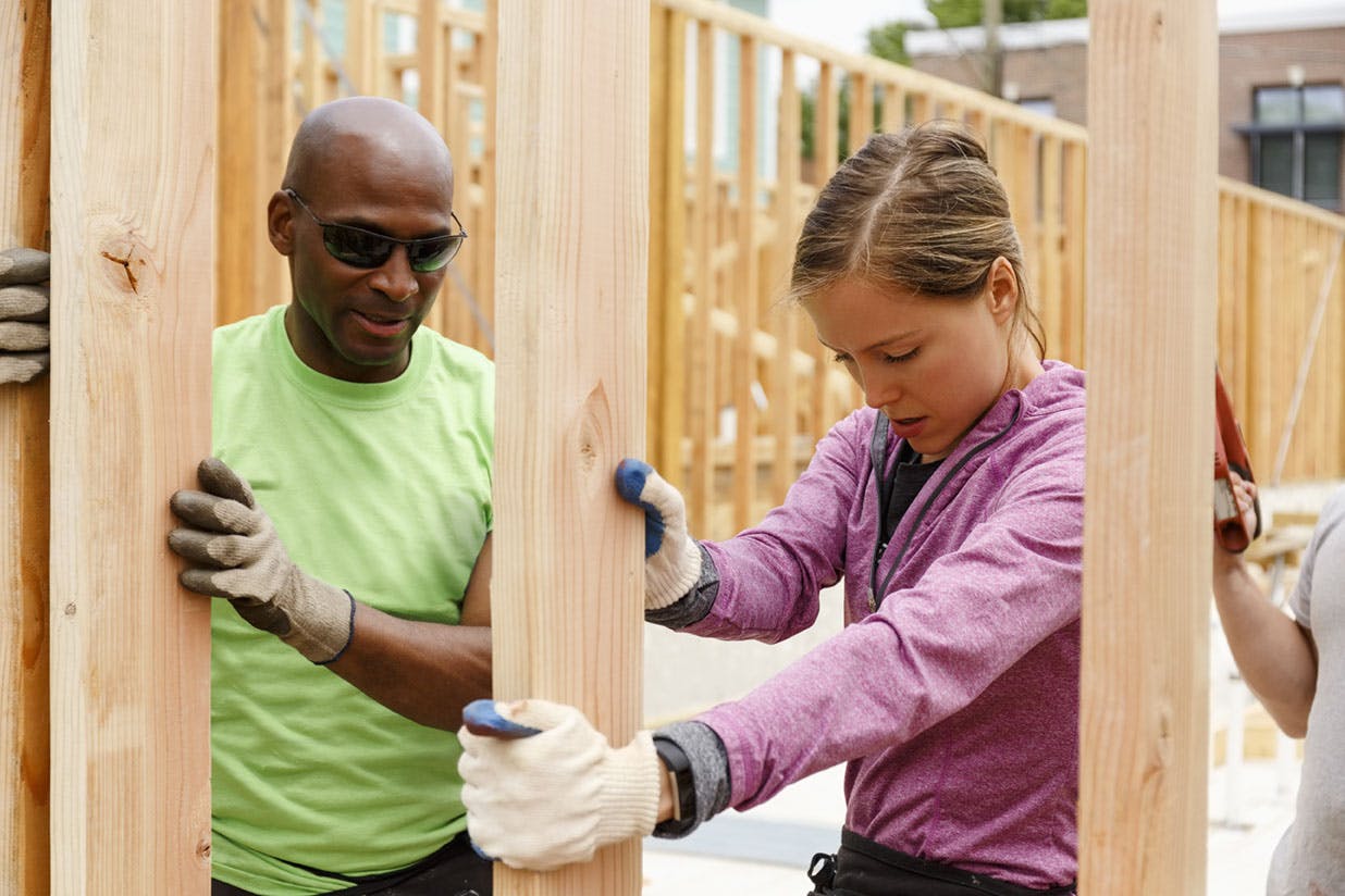 Two people are building a wall together. A women in a pink sweater with gloves on is holding a piece of wood upright. An African American man in a lime green shirt is to her left on the next piece of wood, also holding the wood upright with his gloved hands. He is wearing dark sunglasses. There is a hand to the right of the screen holding a saw.