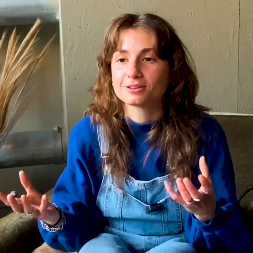 Afbeelding van een vastberaden jongedame van 26 jaar oud. Lang golvend haar, blauwe pull en jeans salopette.