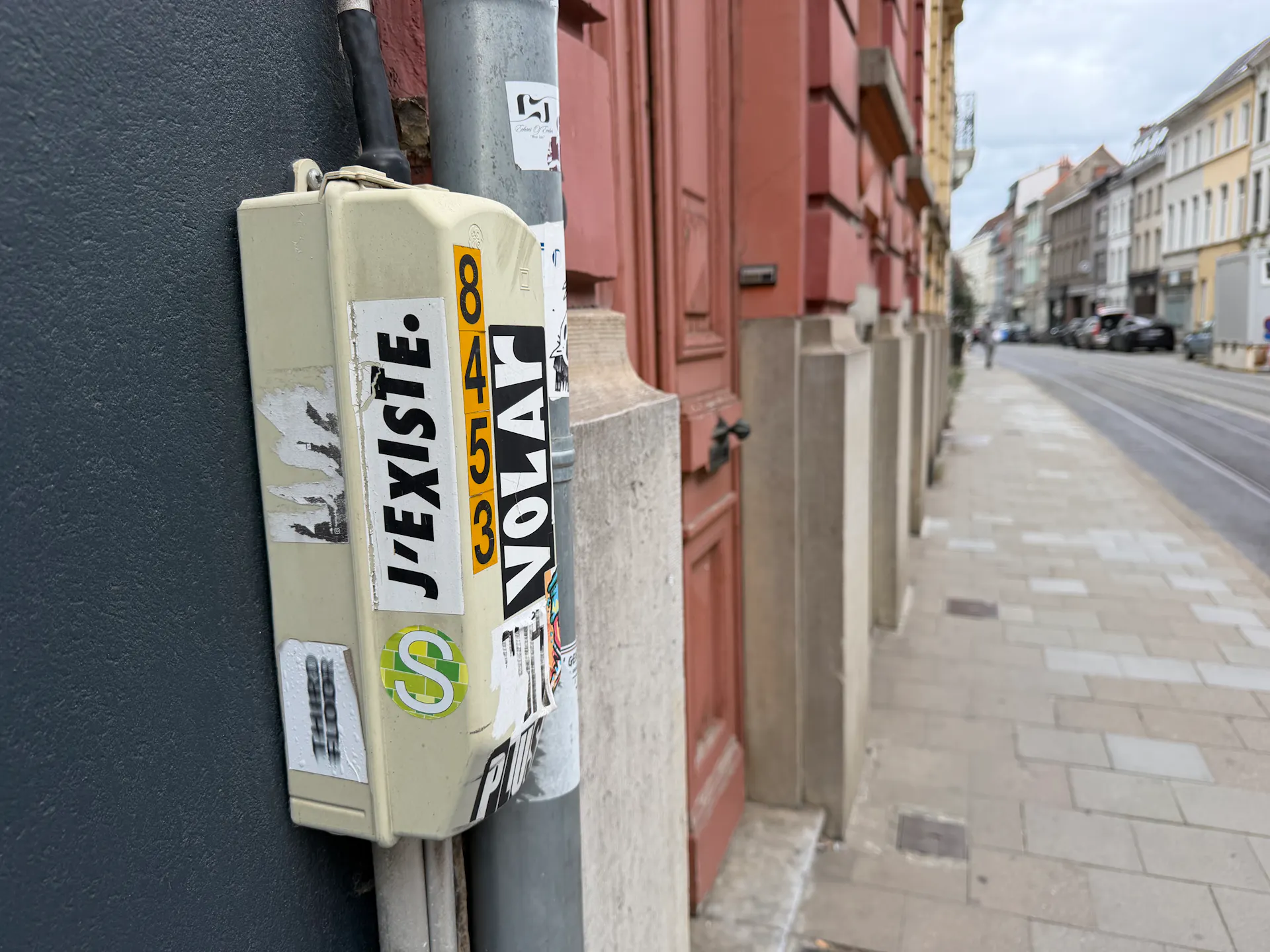 Een beeld van een regenpijp en een doosje dat eraanplakt, waar een sticker opkleeft met de woorden 'J'existe'. In de achtergrond zie je een onscherp beeld van een Gentse straat