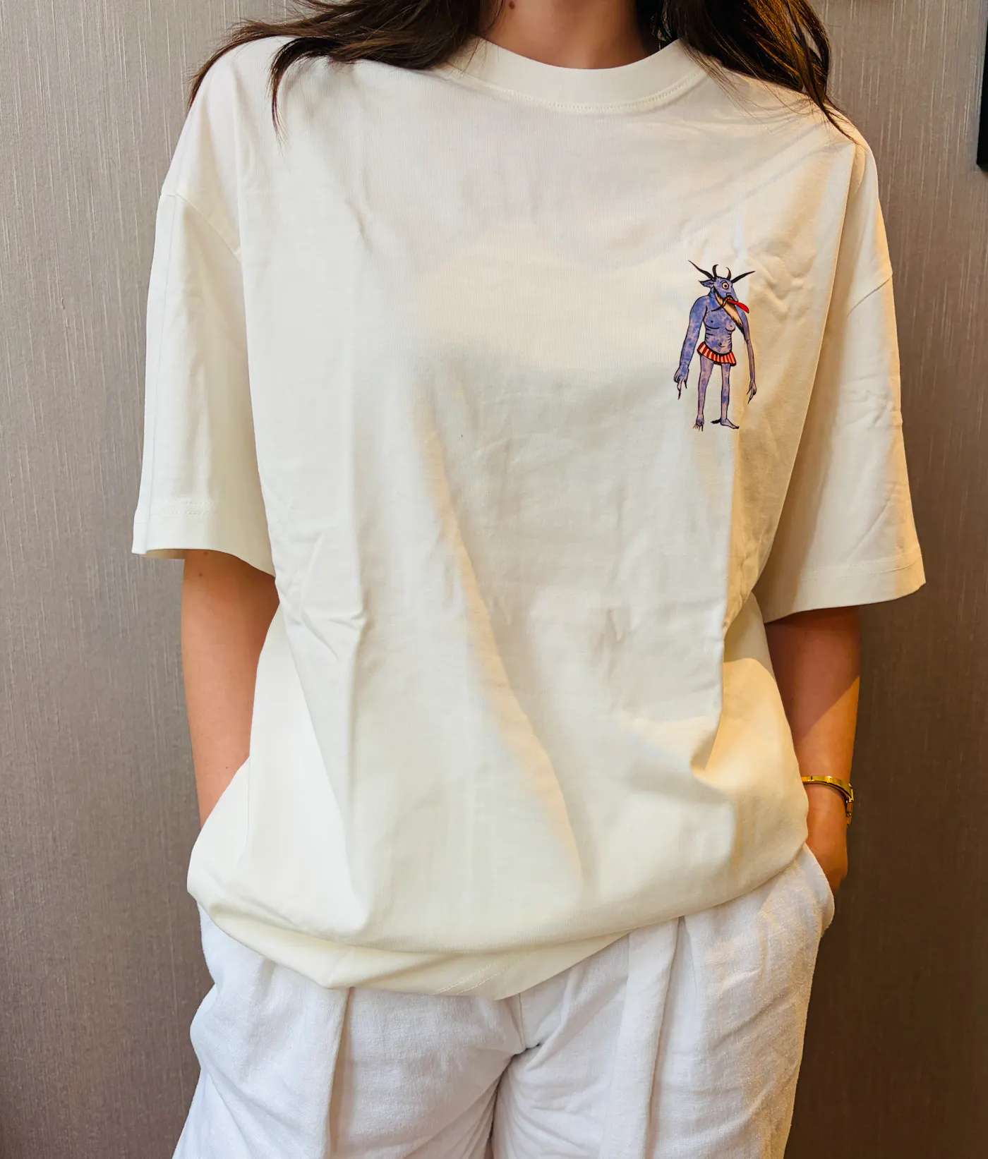 Je ziet een vrouw met een wit t-shirt aan, ze heeft haar handen in de zakken. Je ziet alleen haar bovenlichaam, je ziet niet haar hoofd.