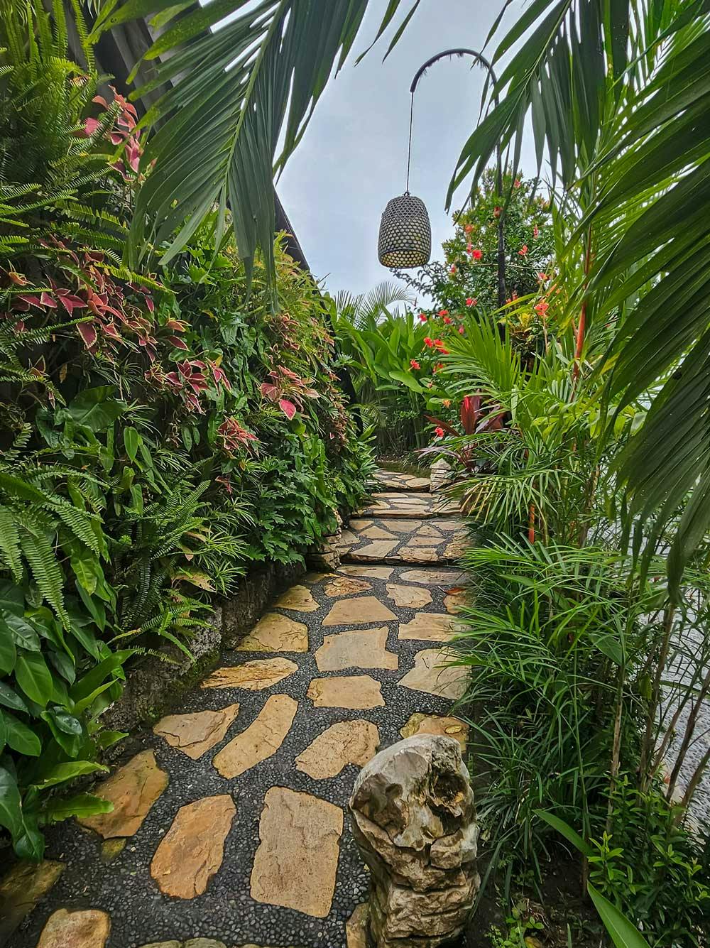 Narrow jungle garden pathway paved with irregular golden stones, lined with ferns, coleus, palms, and red flowering plants, with a woven bamboo pendant lantern overhead