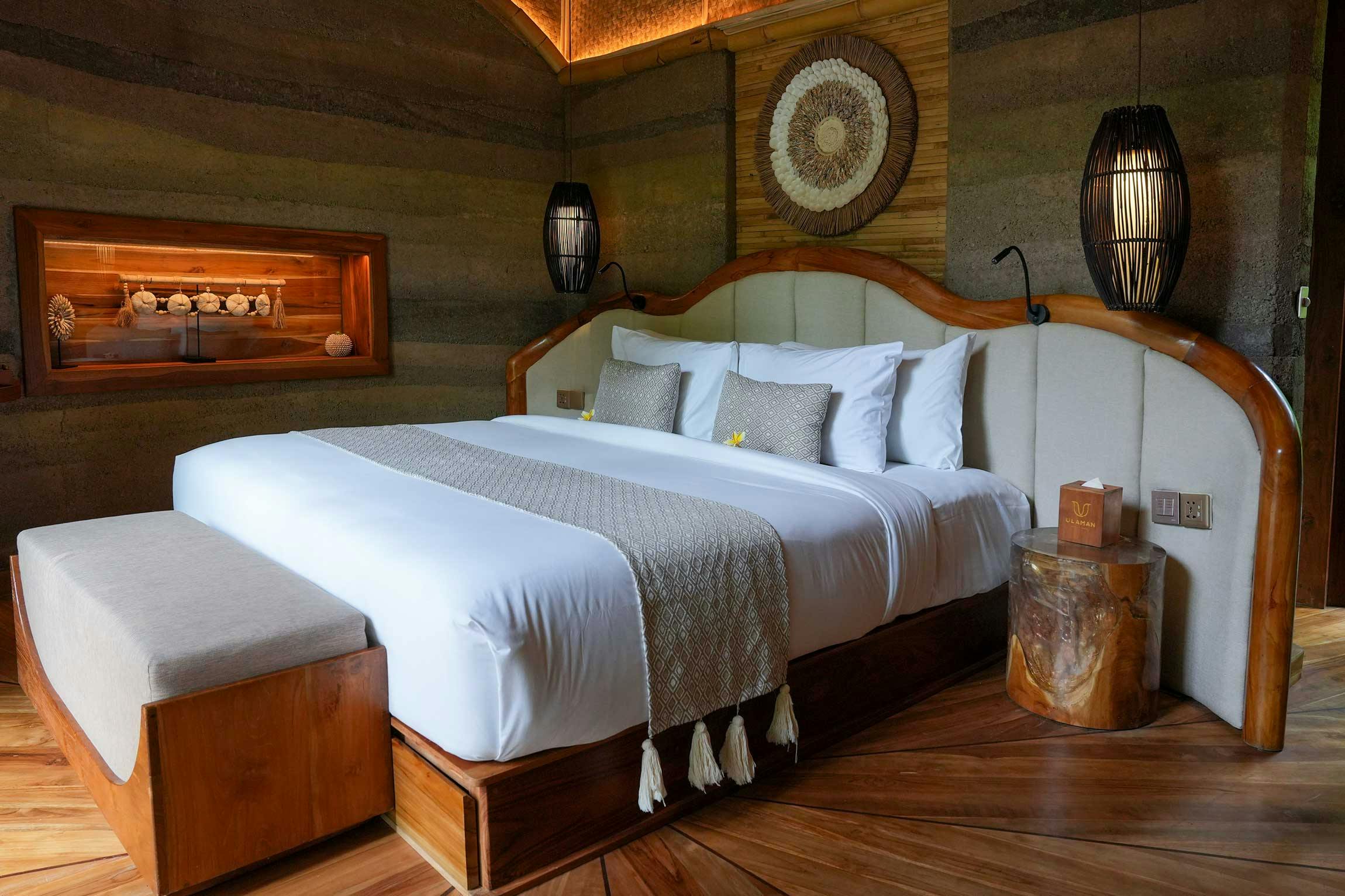 Villa bedroom with a curved teak-framed king bed, woven rattan wall medallion, backlit bamboo ceiling, rammed earth walls, and starburst hardwood floor