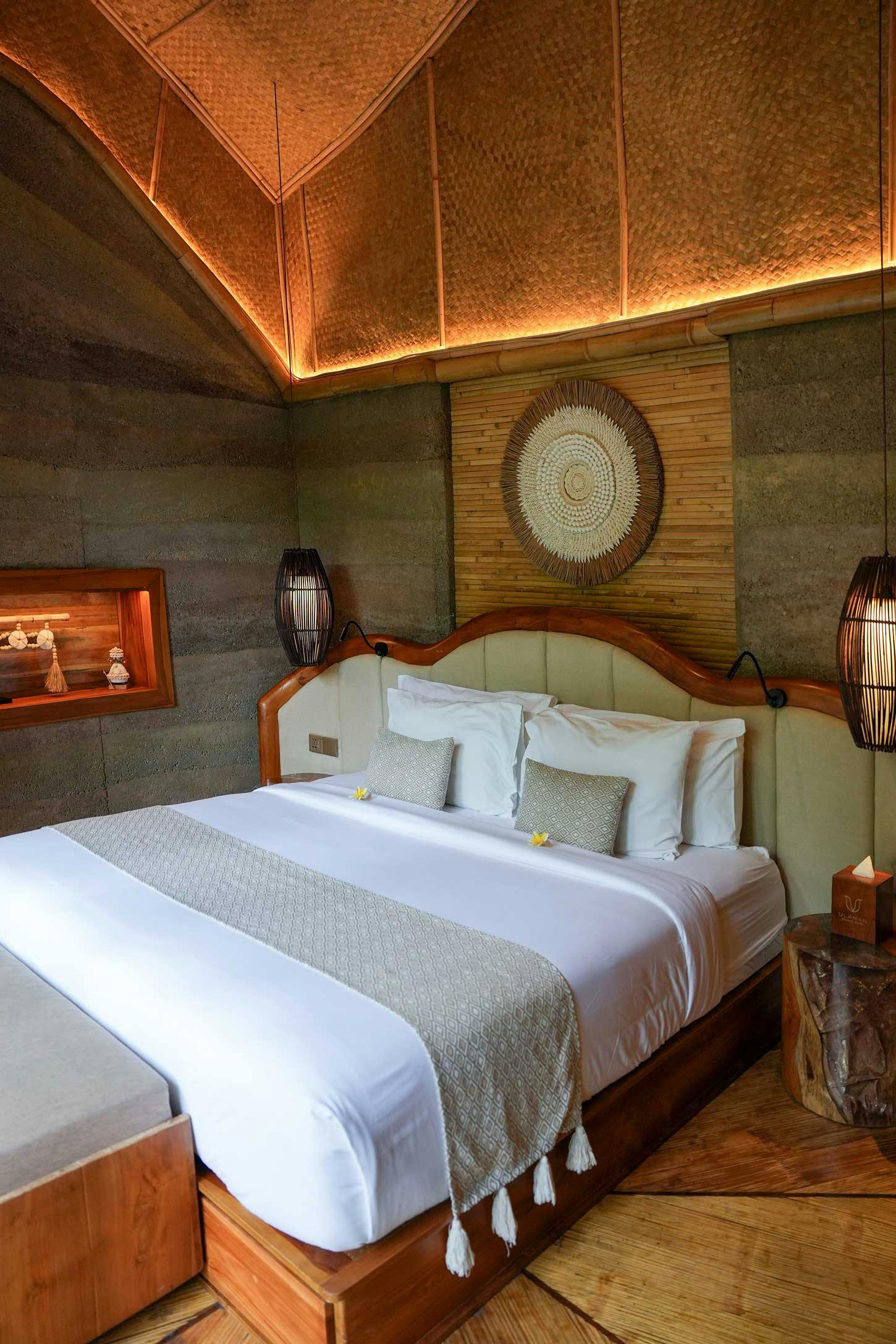 Villa bedroom with a curved teak-framed king bed, woven rattan wall medallion, backlit bamboo ceiling, rammed earth walls, and starburst hardwood floor