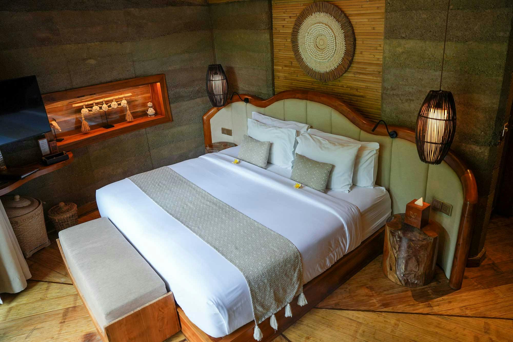 Villa bedroom with a curved teak-framed king bed, woven rattan wall medallion, backlit bamboo ceiling, rammed earth walls, and starburst hardwood floor