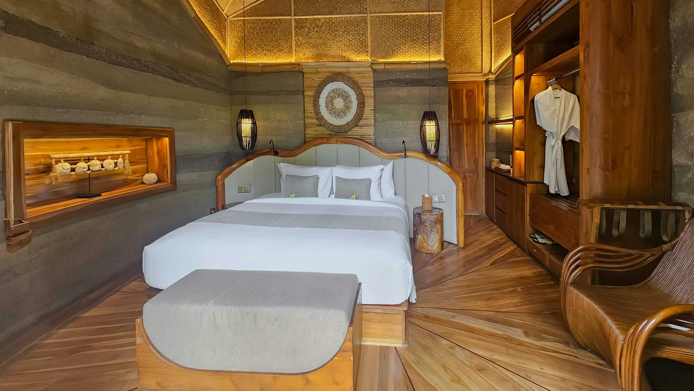 Villa bedroom with a curved teak-framed king bed, woven rattan wall medallion, backlit bamboo ceiling, rammed earth walls, and starburst hardwood floor
