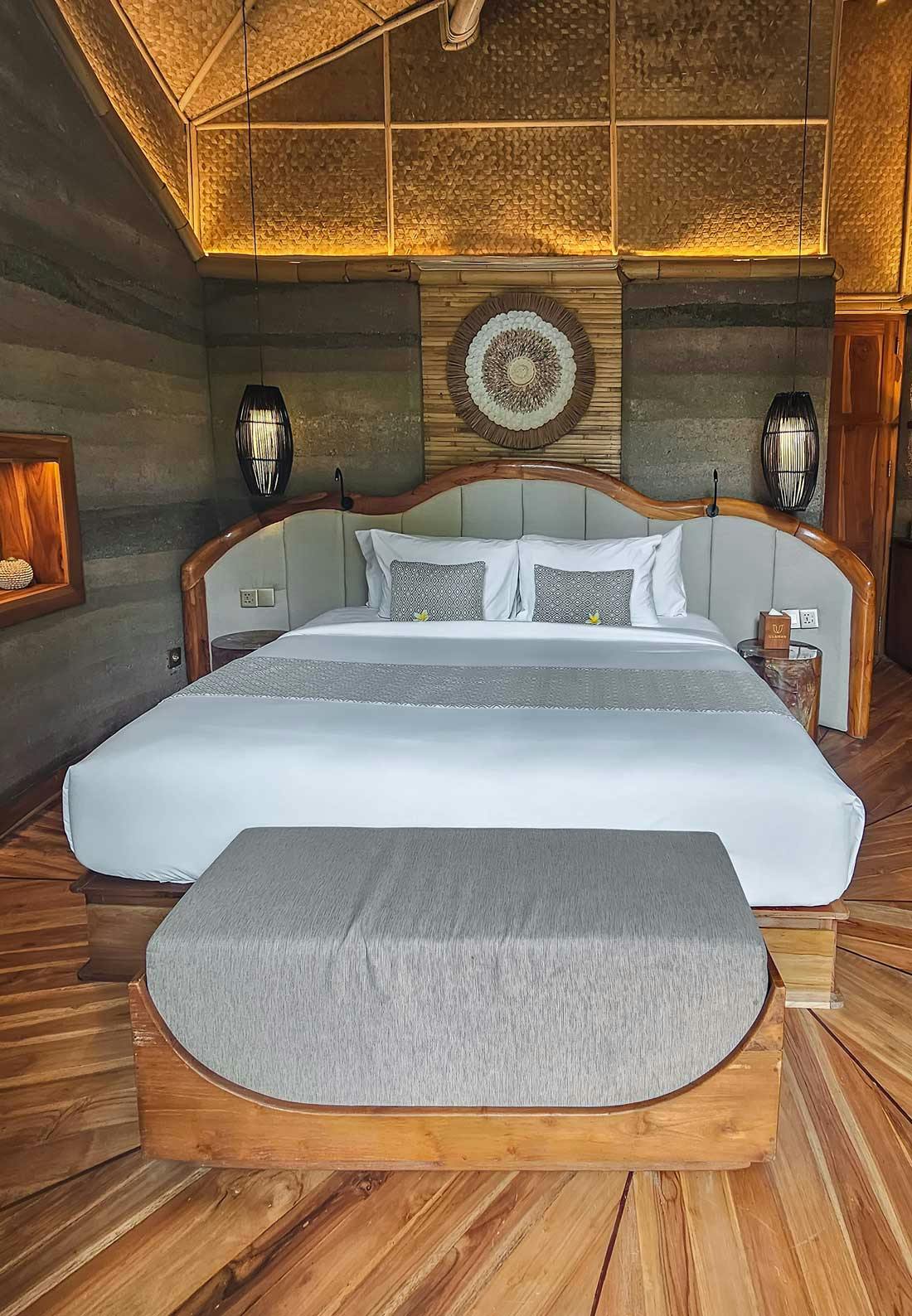 Villa bedroom with a curved teak-framed king bed, woven rattan wall medallion, backlit bamboo ceiling, rammed earth walls, and starburst hardwood floor