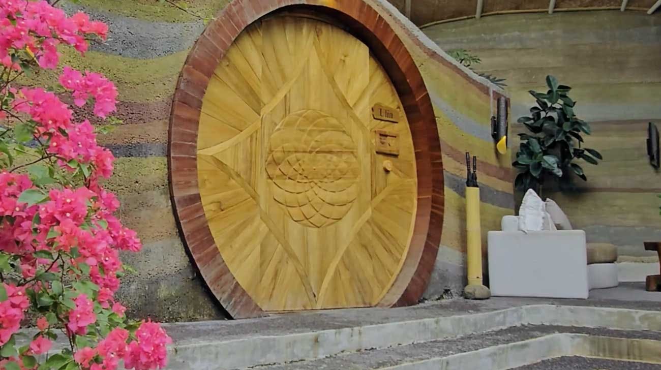 Exterior of the Ulin villa featuring a large circular carved teak door set into rammed earth walls, framed by vibrant pink bougainvillea blooms and a bamboo-structured roof above
