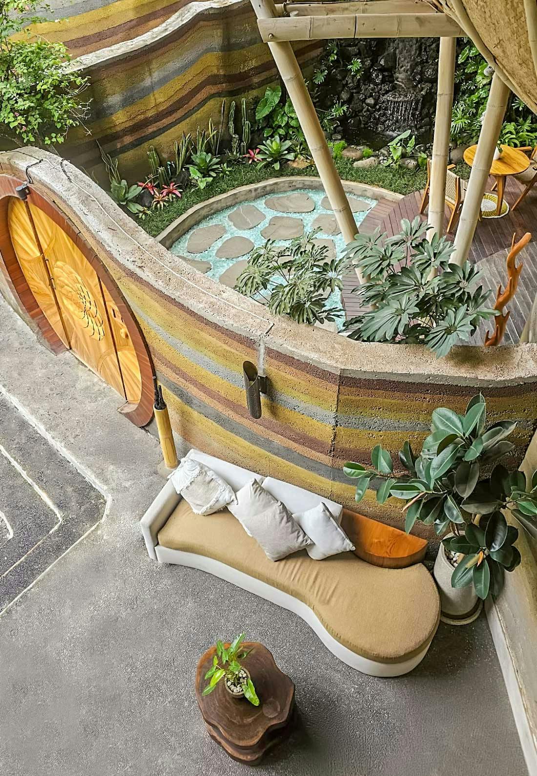 Aerial view of villa entrance with a circular carved wooden door, rammed earth walls, with stepping stones, and a curved beige sofa on a concrete terrace