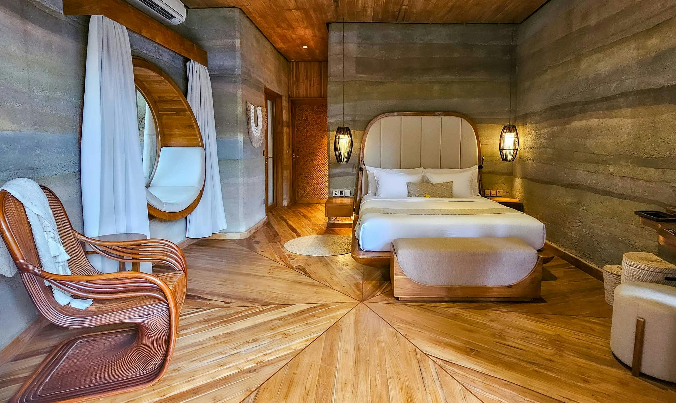 Warm villa bedroom interior with a curved upholstered headboard, starburst-patterned hardwood floor, rammed earth walls, rattan chair, and an oval porthole window with white drapes