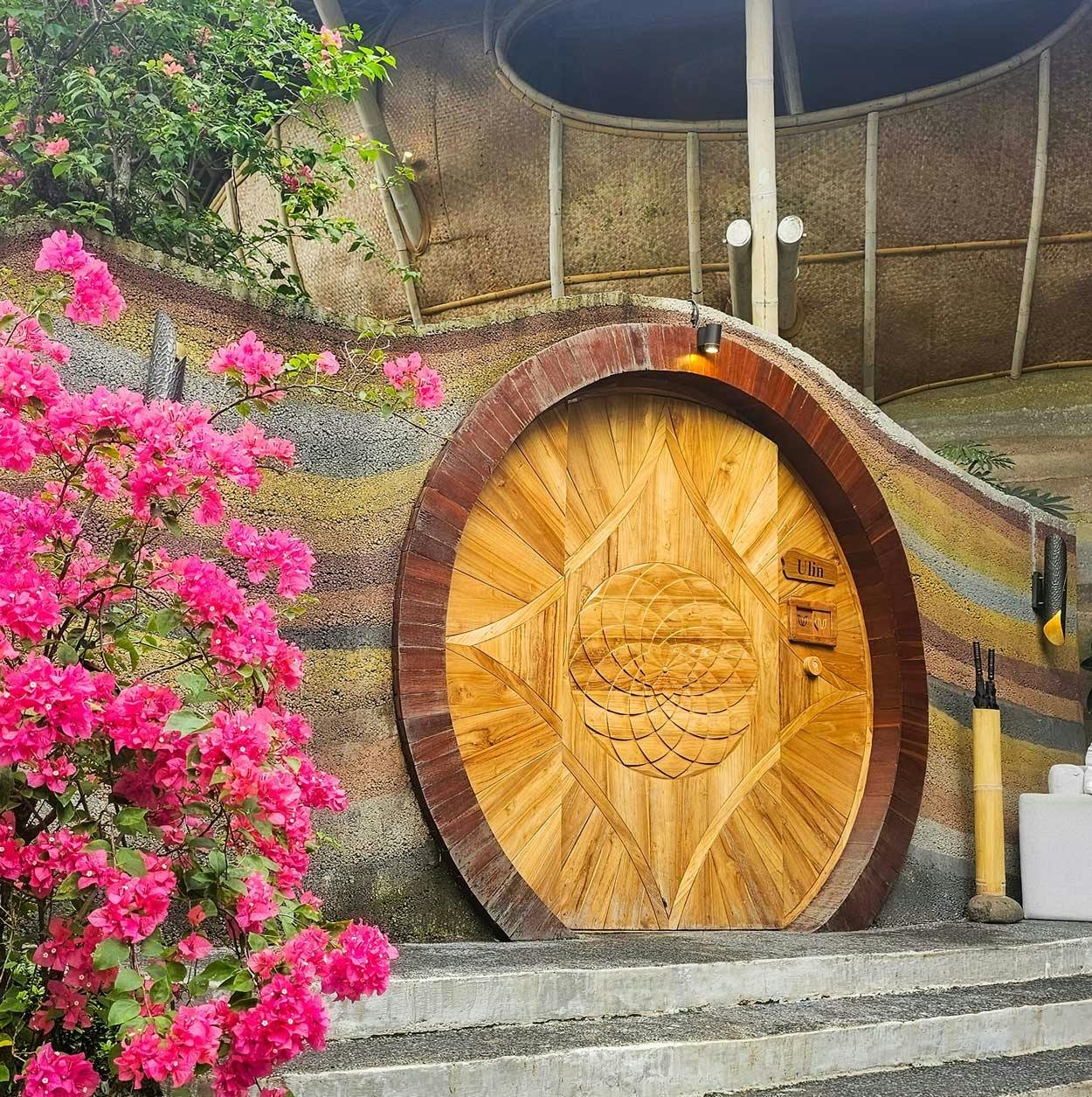 Exterior of the Ulin villa featuring a large circular carved teak door set into rammed earth walls, framed by vibrant pink bougainvillea blooms and a bamboo-structured roof above