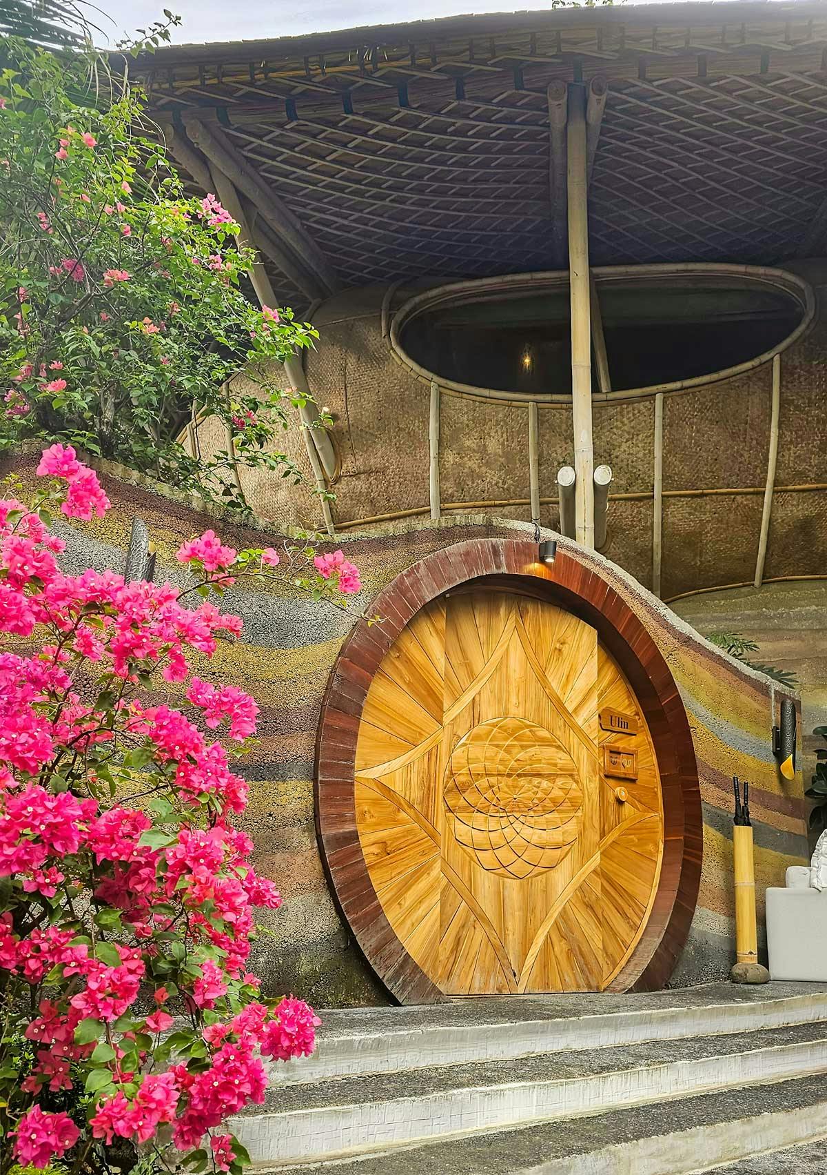 Exterior of the Ulin villa featuring a large circular carved teak door set into rammed earth walls, framed by vibrant pink bougainvillea blooms and a bamboo-structured roof above