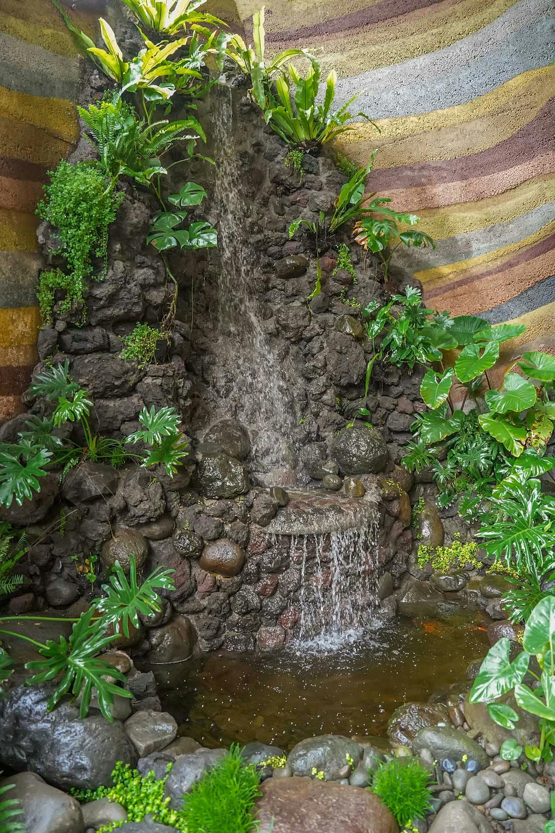A tiered stone waterfall cascading into a small koi pond, surrounded by tropical ferns, monstera, and leaves against the villa's signature rammed earth walls