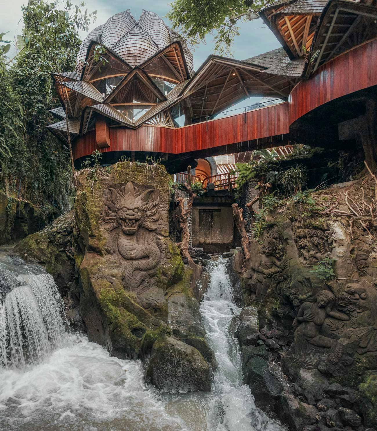 Dramatic resort architecture featuring a lotus-shaped wooden dome and angular thatched rooftops rising above a rushing waterfall, with intricate stone dragon carvings and sculptures carved into boulders in a lush jungle ravine.