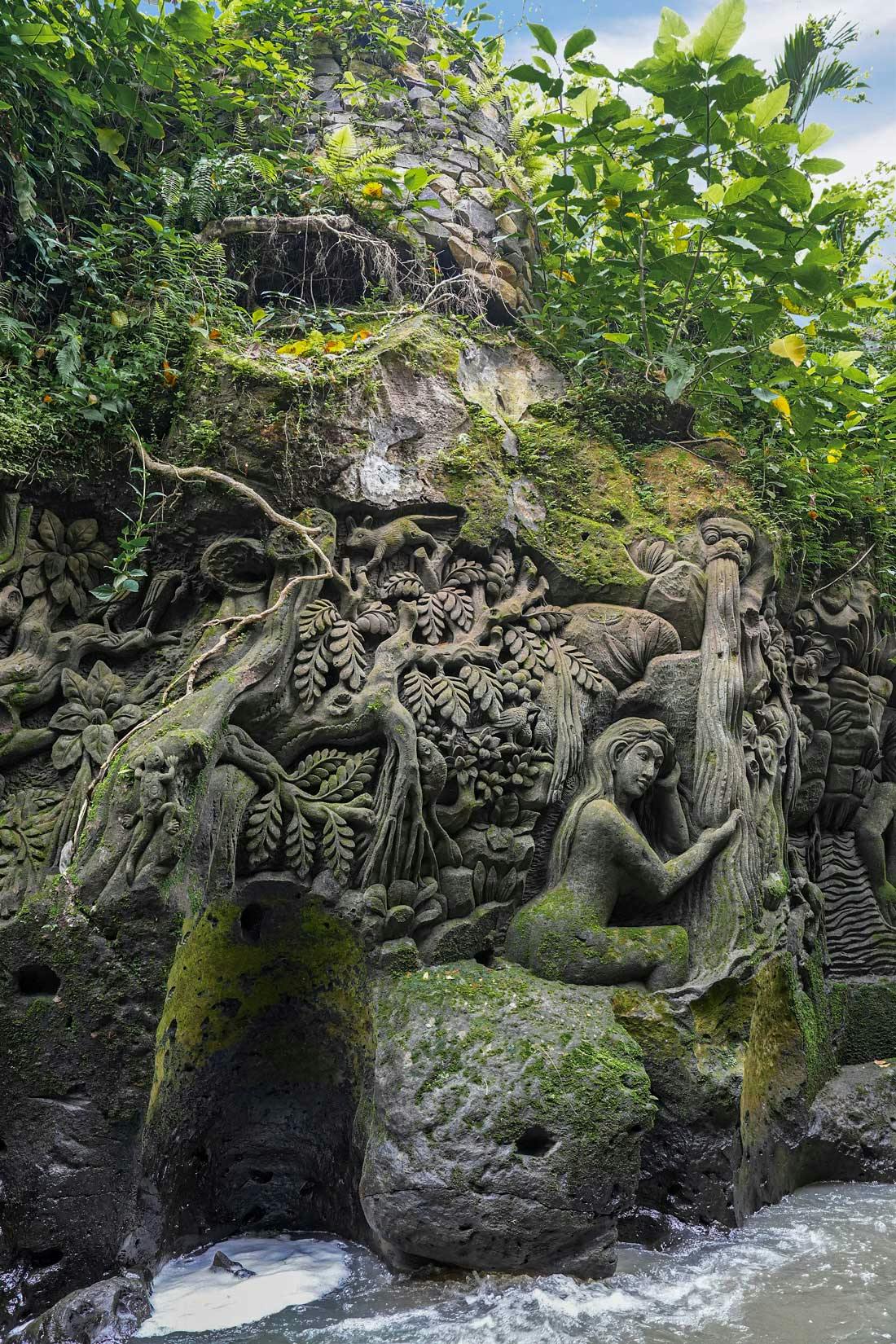 Detailed Balinese stone relief carvings surrounded by intricate foliage, animals, and floral motifs in a dense tropical jungle setting.