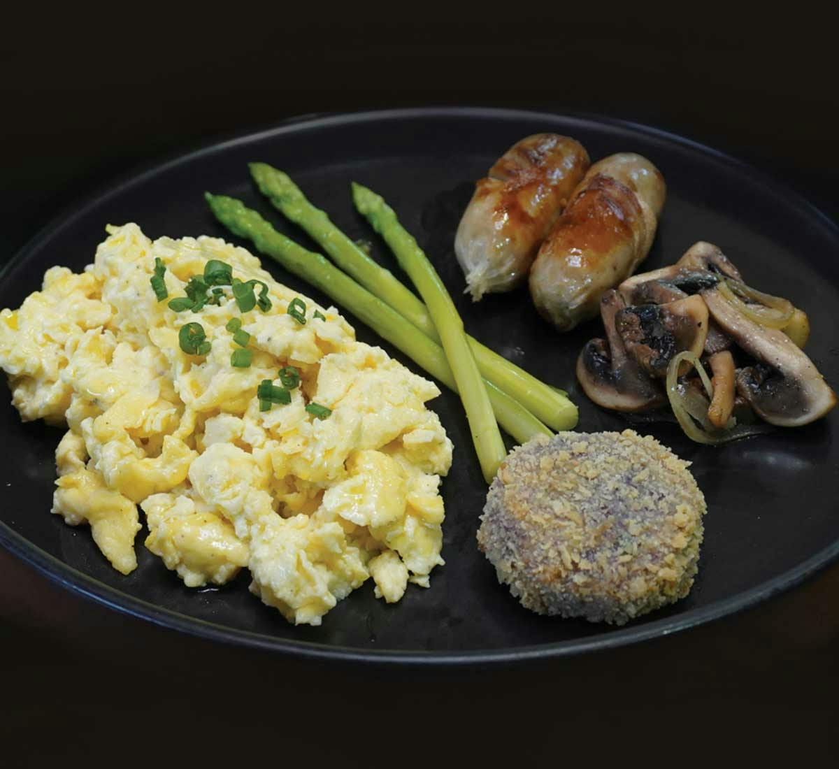 Scrambled eggs with asparagus, chicken sausages, sautéed mushrooms, and a sweet potato fritter on a black plate.