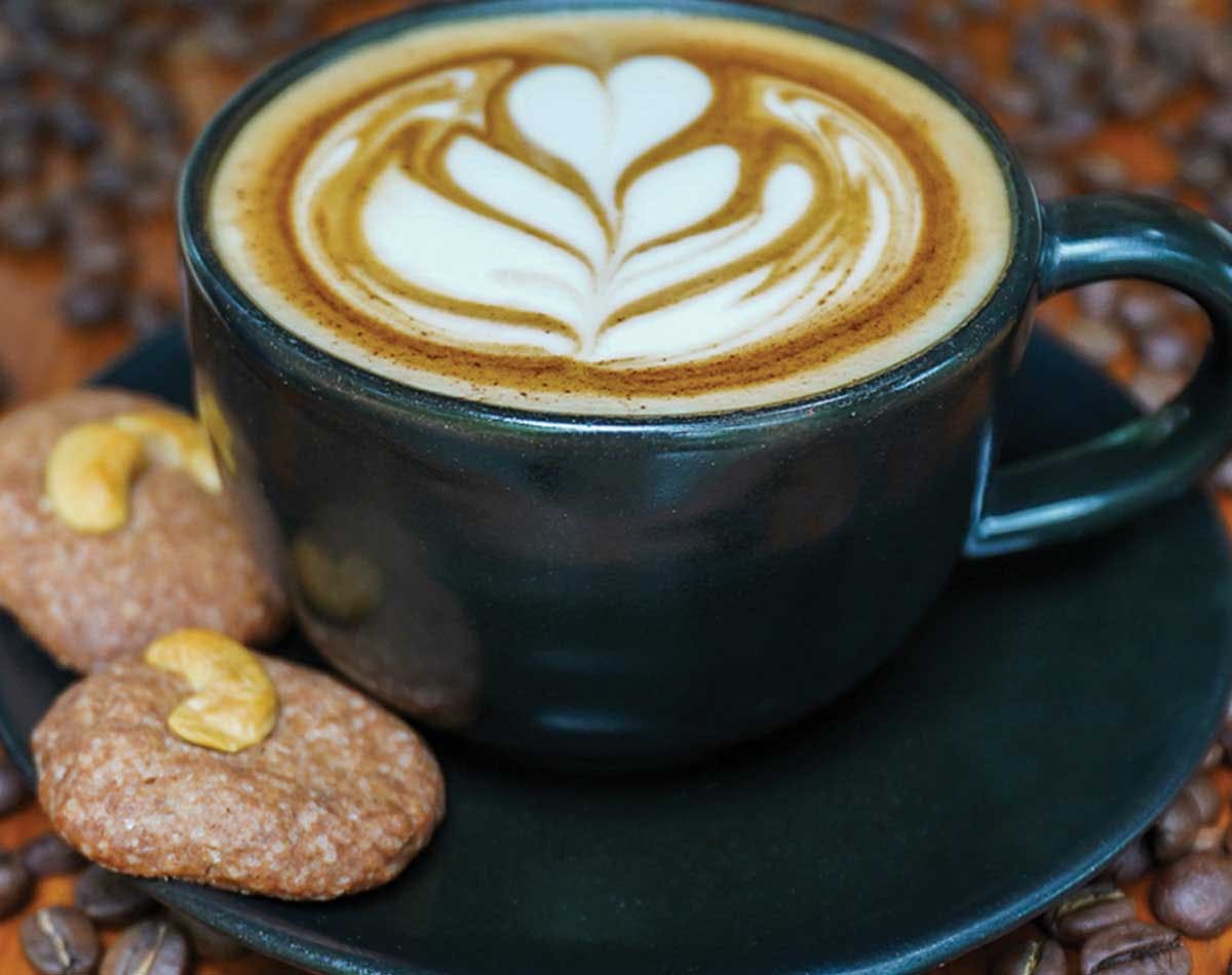 Latte art in a dark green ceramic cup, surrounded by coffee beans and cashew cookies on a saucer.