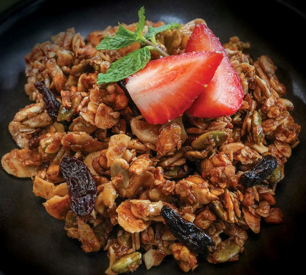 Close-up of homemade granola with dried fruit and seeds in a black bowl, topped with a sliced strawberry and fresh mint.