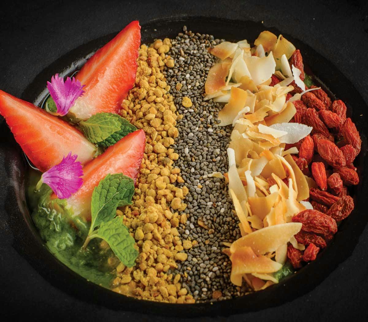 Bliss smoothie bowl with neat rows of bee pollen, chia seeds, coconut flakes, and goji berries, garnished with fresh strawberries and edible flowers.
