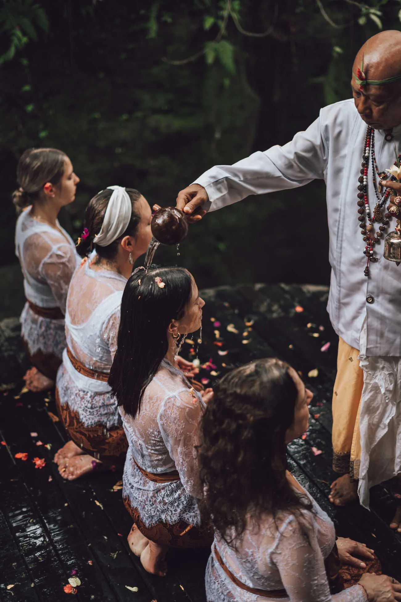 Balinese Hindu priest performing a traditional water blessing ceremony over retreat guests surrounded by flower petals at a sacred jungle retreat in Bali