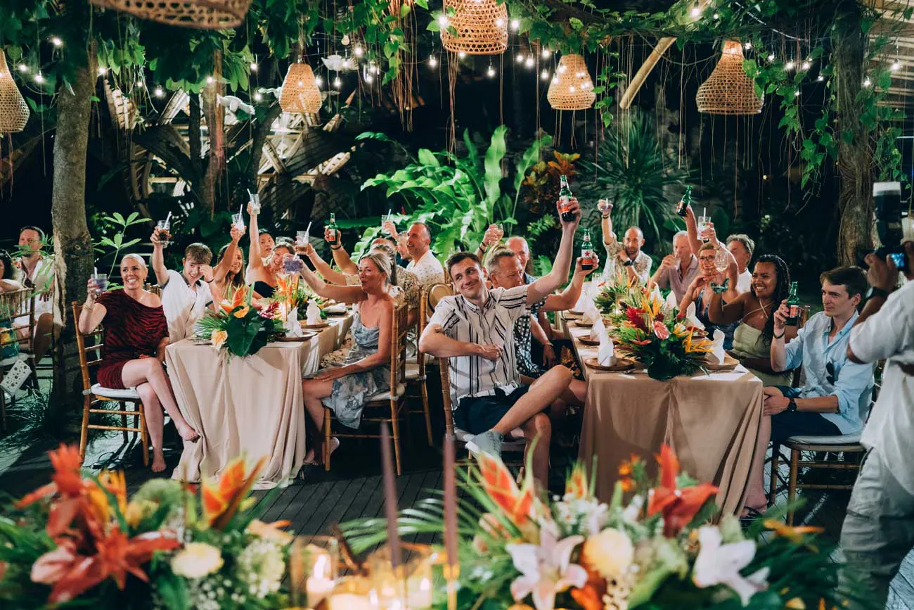 Wedding guests raising glasses in a toast under woven rattan pendant lights and tropical foliage at a jungle wedding reception in Bali