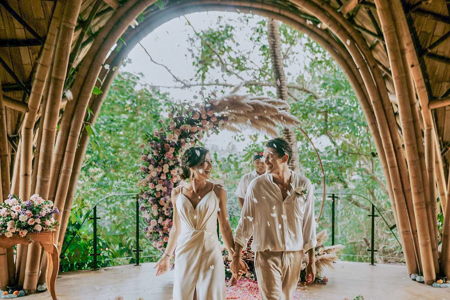 Bride and groom walking hand in hand through a stunning bamboo arch ceremony space with colorful floral arrangements and confetti at a Bali wedding