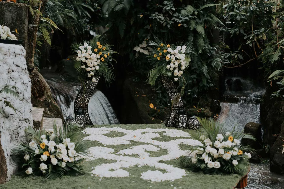Elegant outdoor wedding ceremony setup with white floral arrangements, petal-lined aisle, and cascading waterfalls at a lush Ubud Bali wedding venue