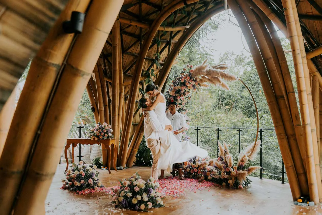 Couple sharing their first kiss at Bali wedding ceremony inside an arched bamboo shala with a colorful circular floral arch and petal-strewn floor