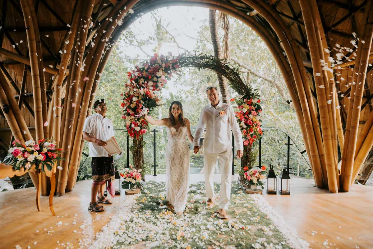 Newlyweds walking down the aisle beneath falling flower petals inside a bamboo arch ceremony space with vibrant tropical floral arrangements at a Bali wedding venue