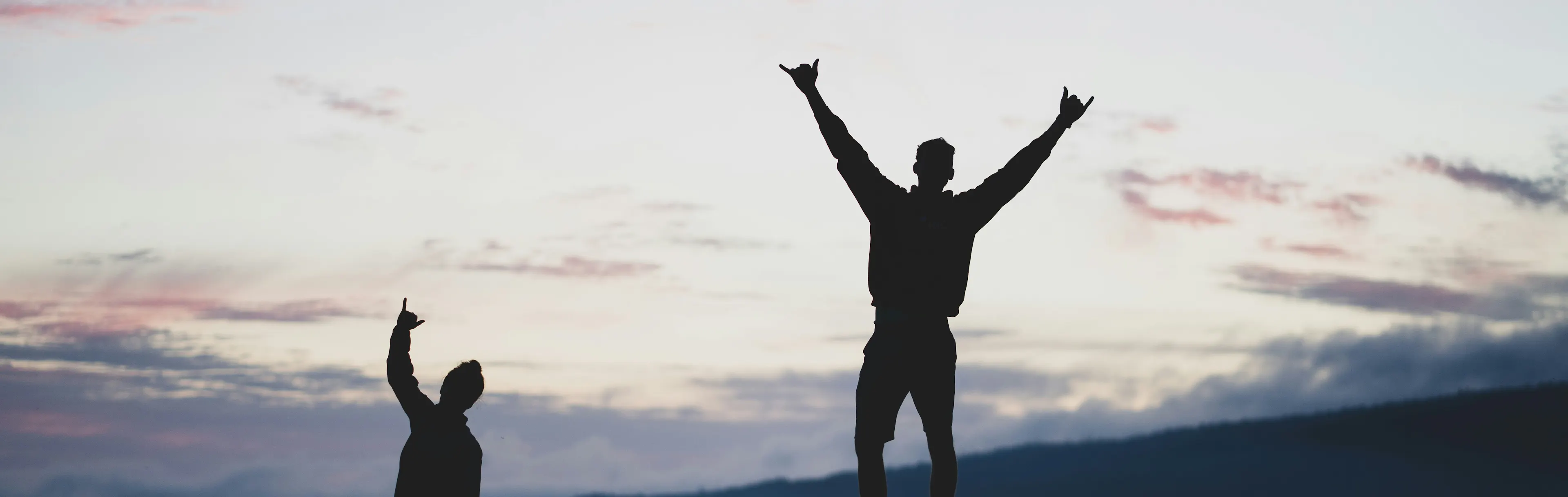 Two people silhouetted on rocks at sunset with arms raised in celebration
