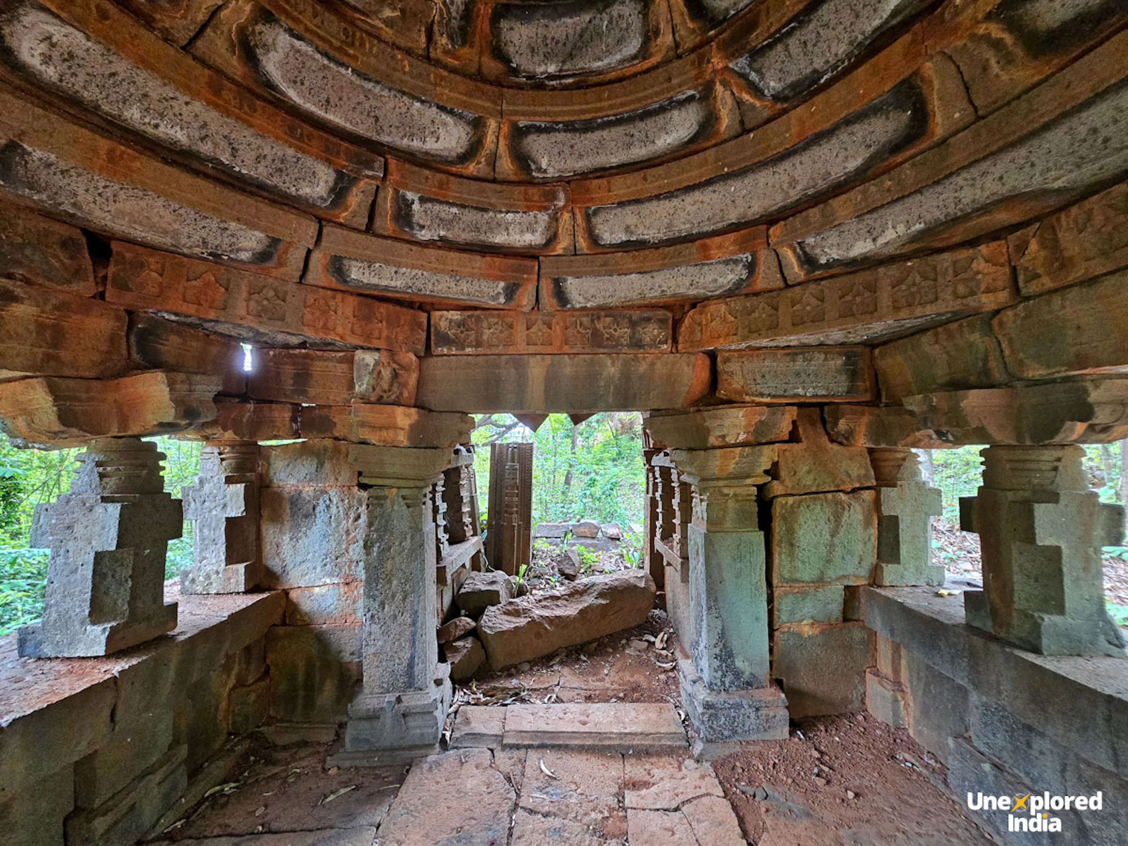 Interior of Sardesai Wada, showing stone pillars, carved walls and a domed ceiling surrounded by greenery.