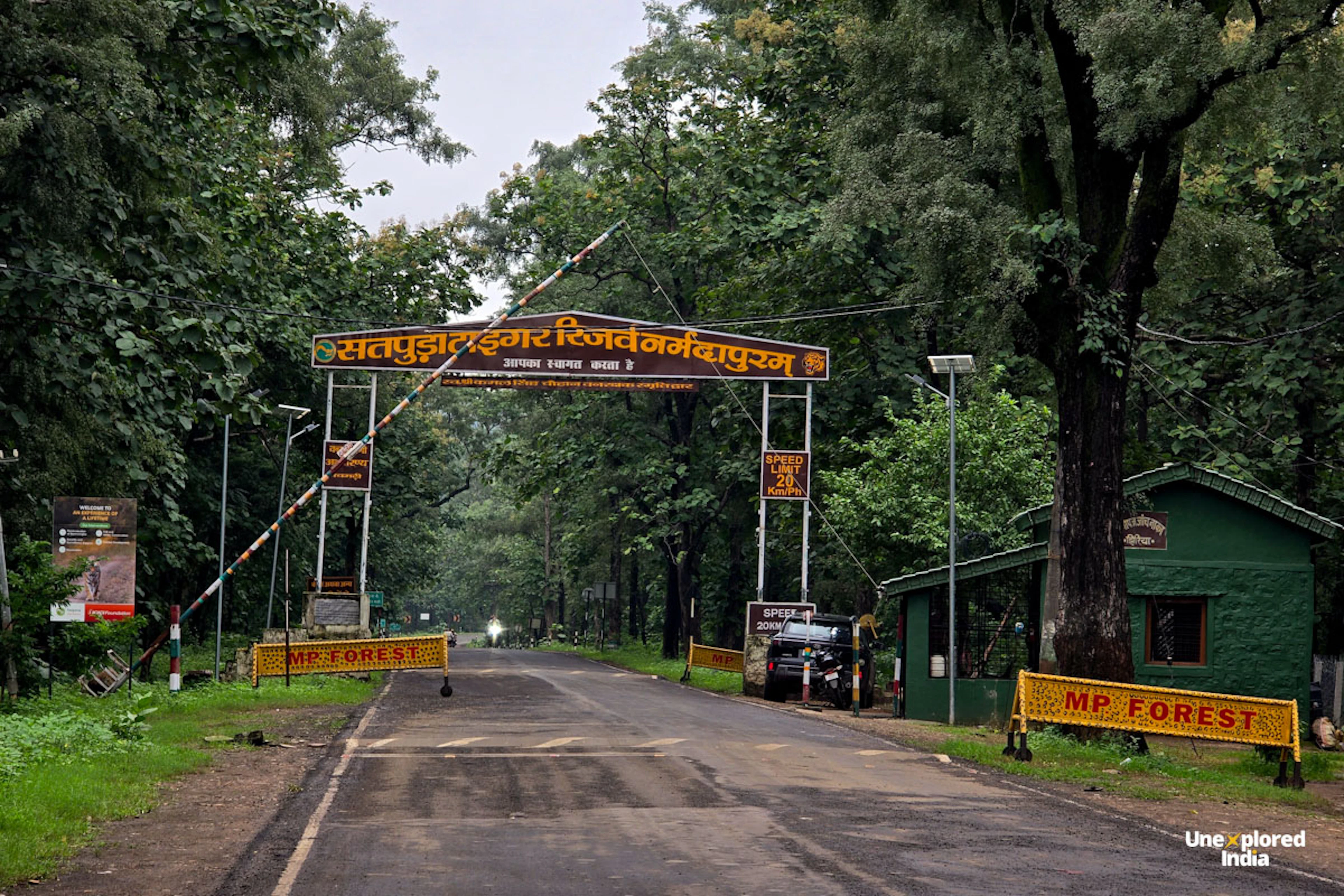 Entrance gate of Satpura Tiger Reserve, with an arched Hindi signboard over a road that leads into dense forest, a raised barrier, yellow “MP Forest” barricades and a green forest checkpost beside the road.