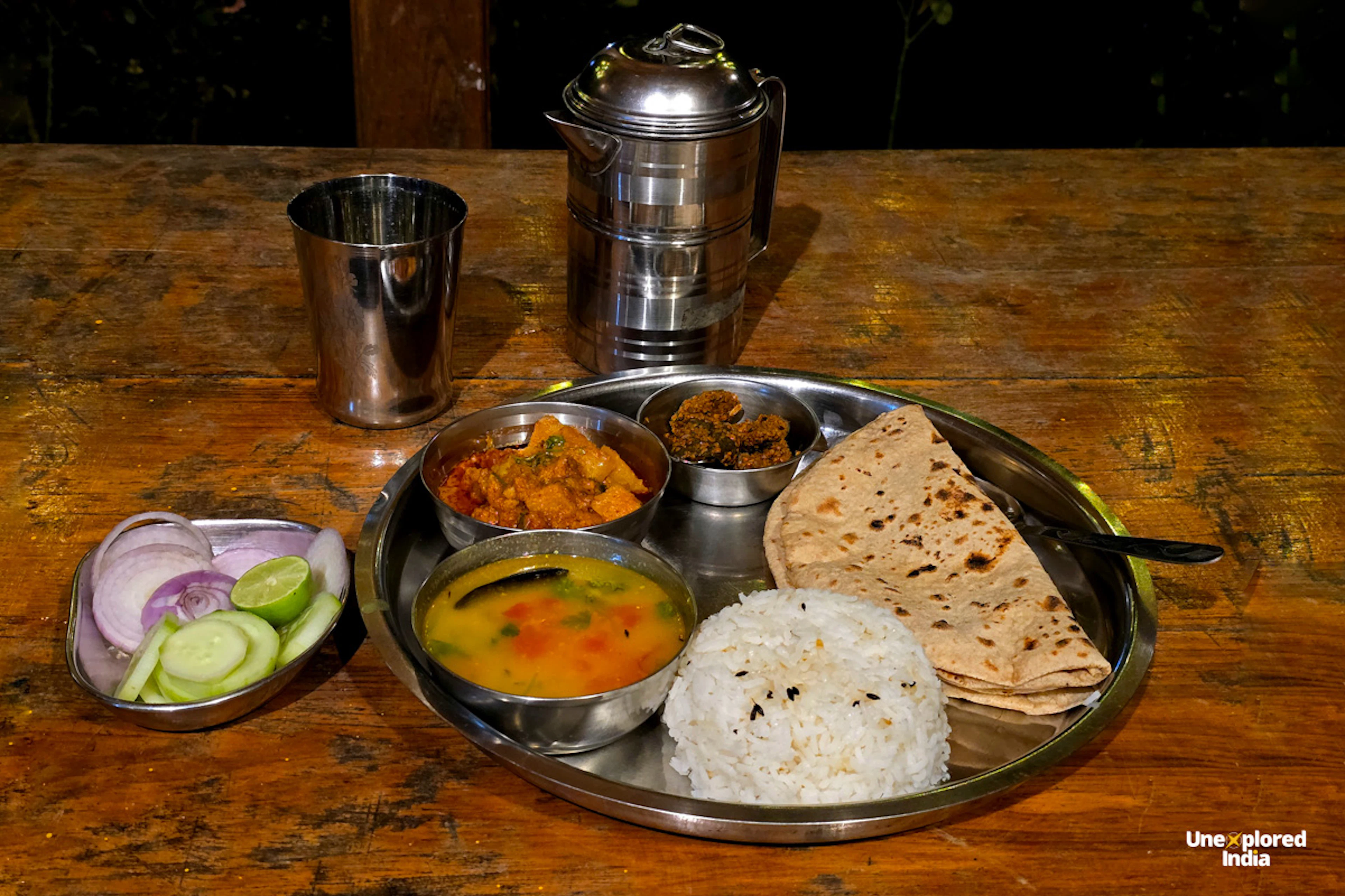 A steel thali on a wooden table with dal, a vegetable curry, a dry sabzi, rice and folded chapatis, beside a side plate of sliced onion, cucumber and lime, a steel tumbler and a tiffin carrier.