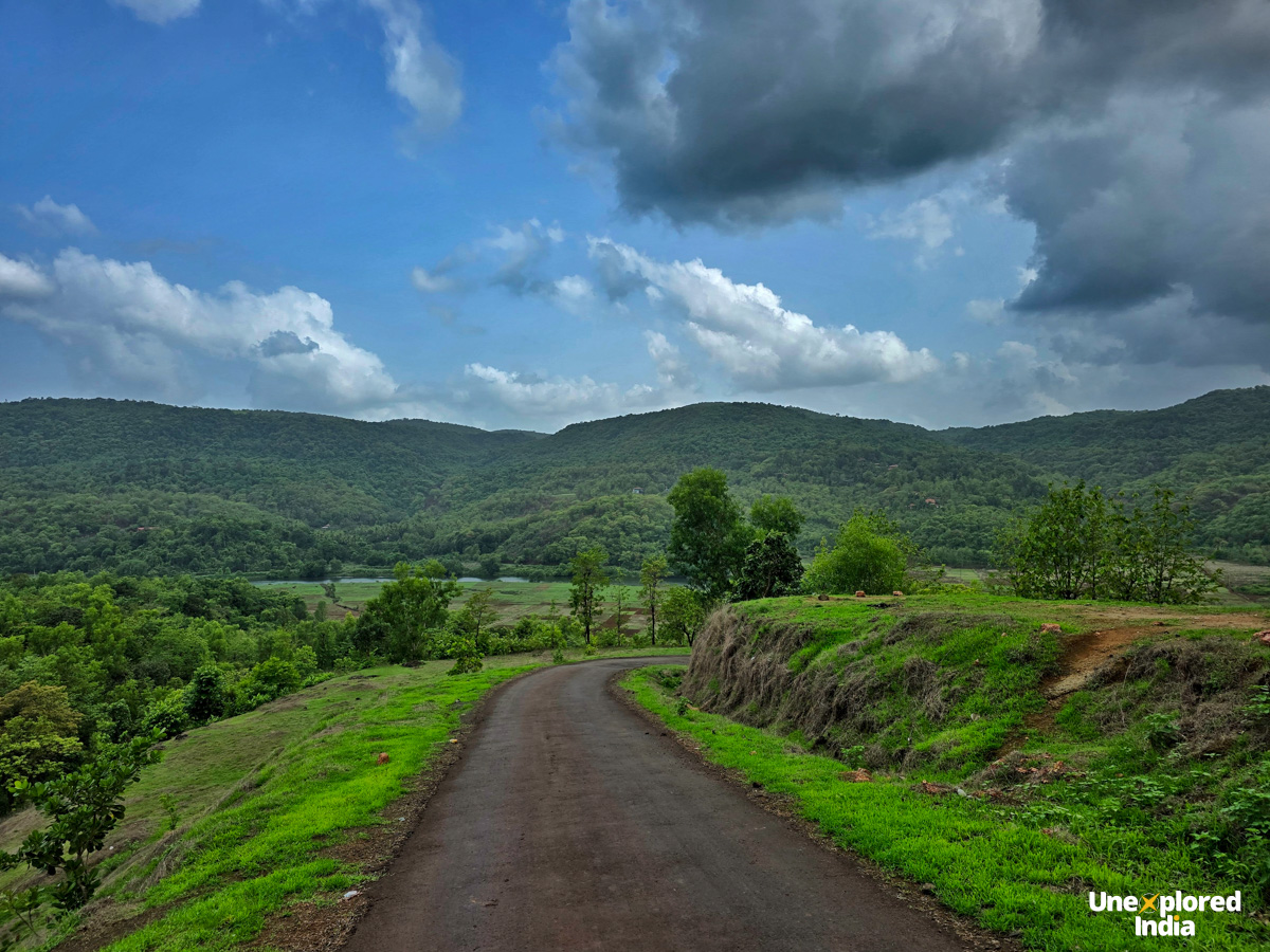 A winding road through the green hills and fields of Tural village in the Konkan region under a cloudy monsoon sky.
