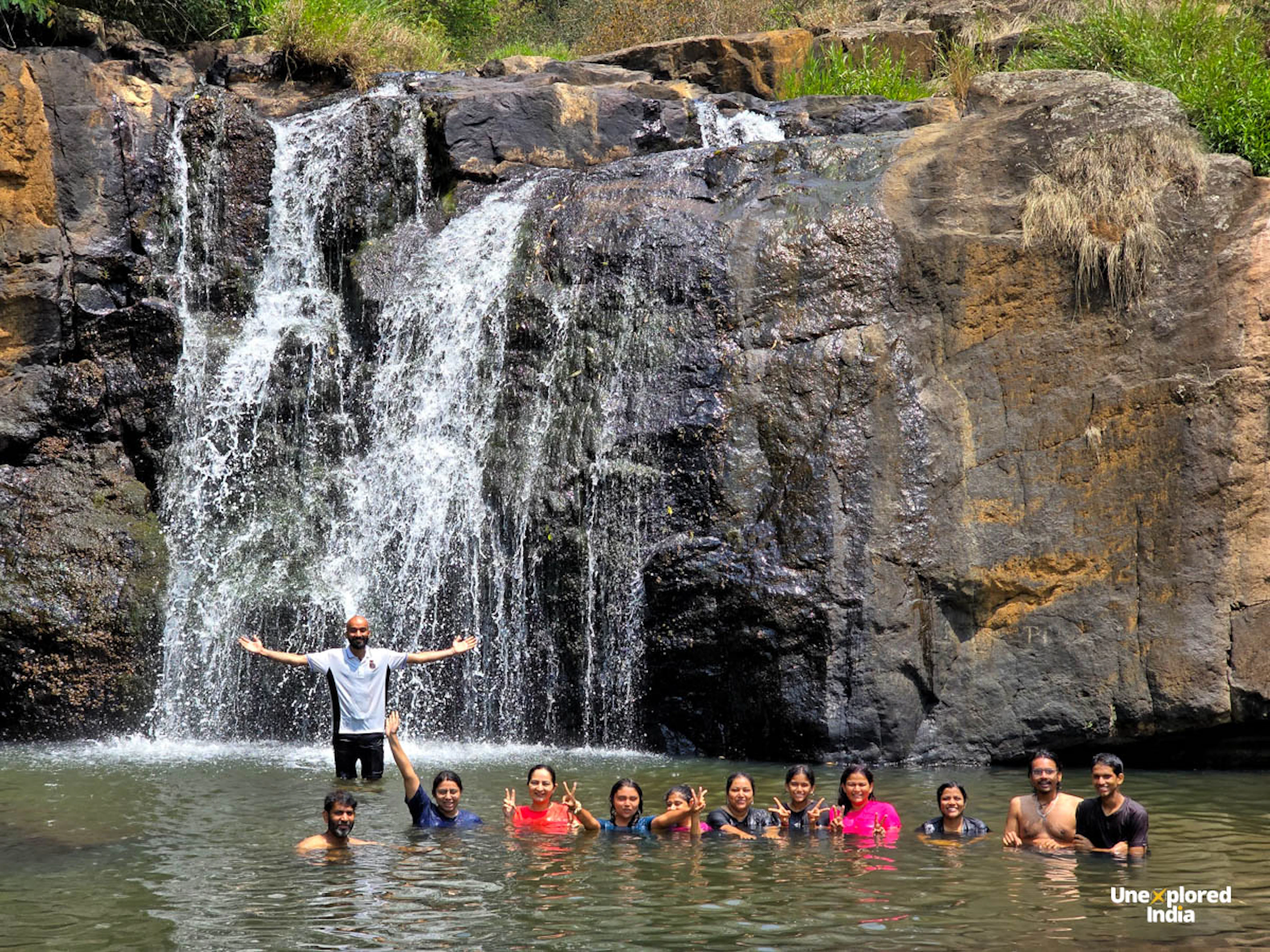 A group swims beneath Kundha waterfalls in the Nilgiris, surrounded by rocky cliffs, flowing water and lush greenery.