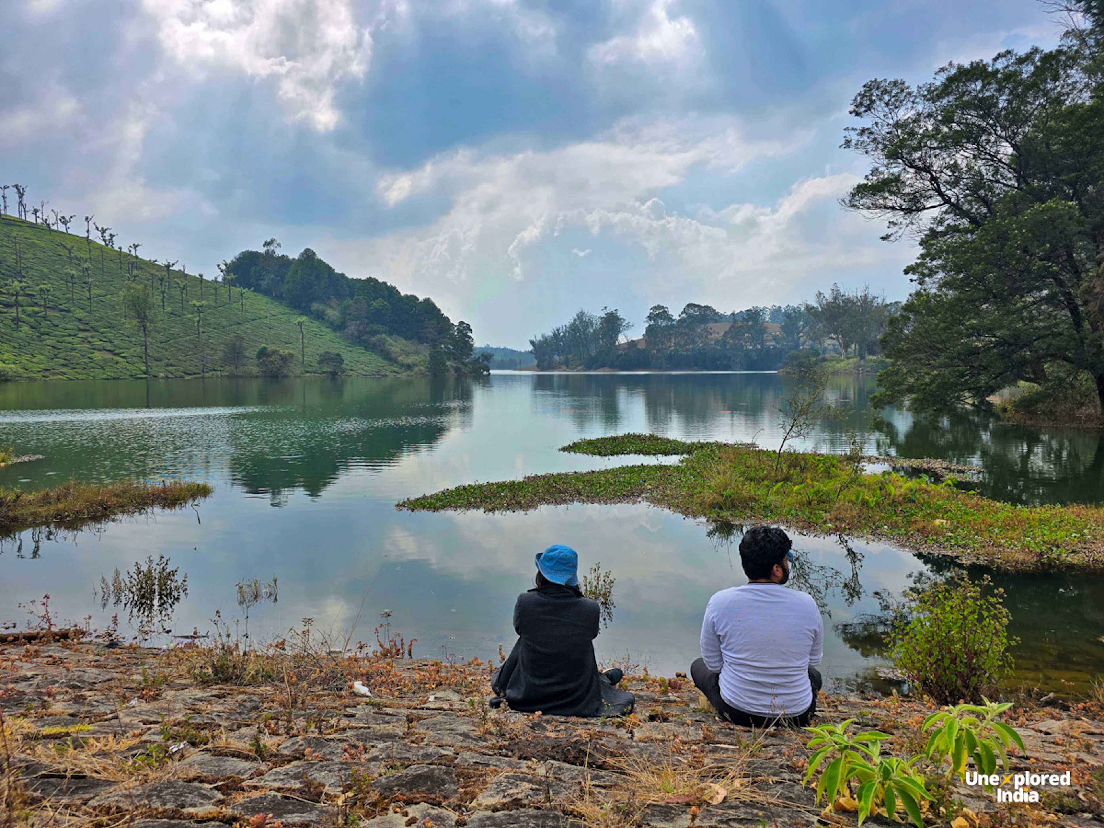 Ooty lake surrounded by tea plantations and hills in the Nilgiris, with two people sitting by the water.