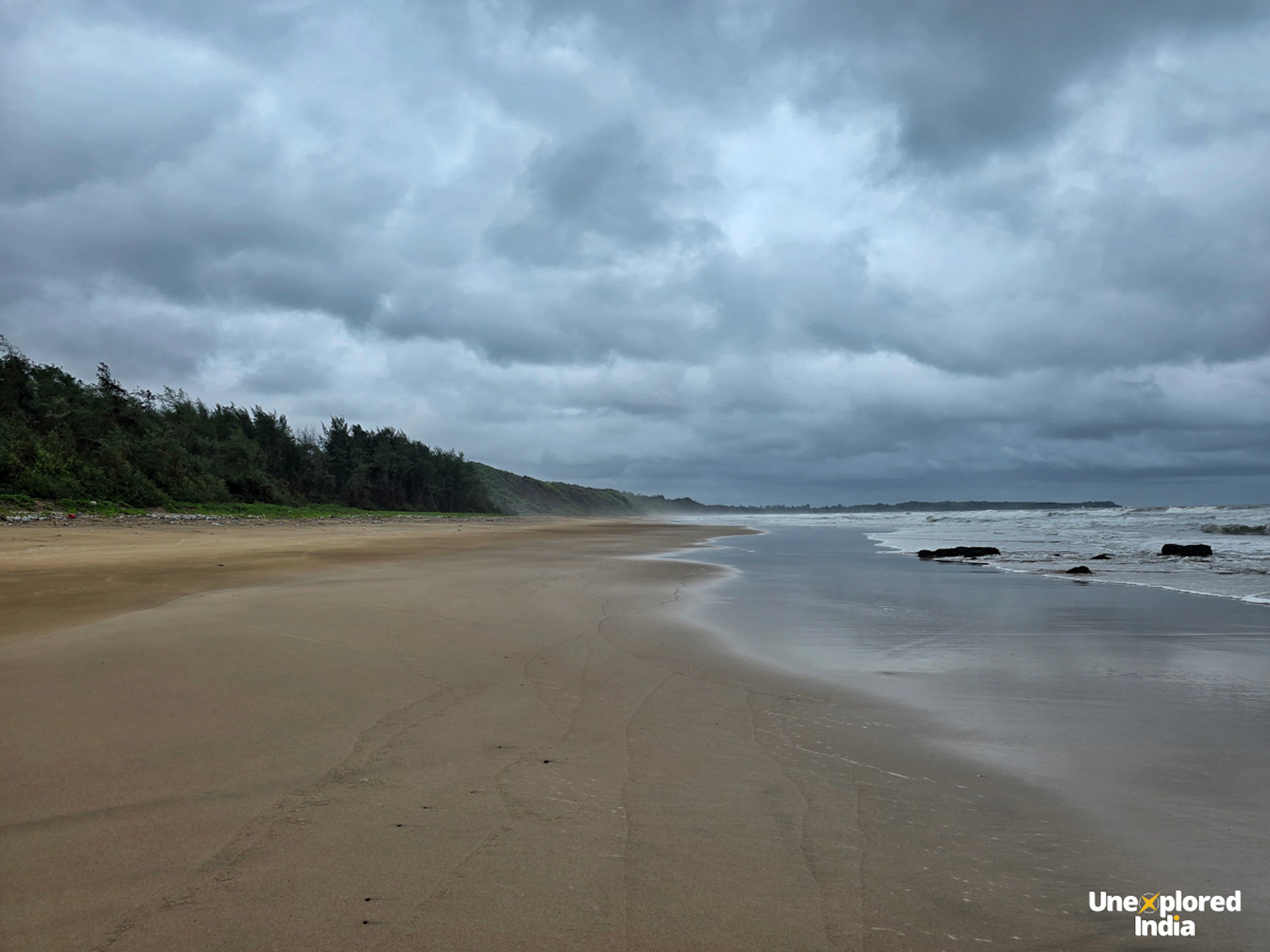 A quiet Konkan beach stretches along the coast under dark monsoon clouds, with waves rolling onto the wide sandy shore.