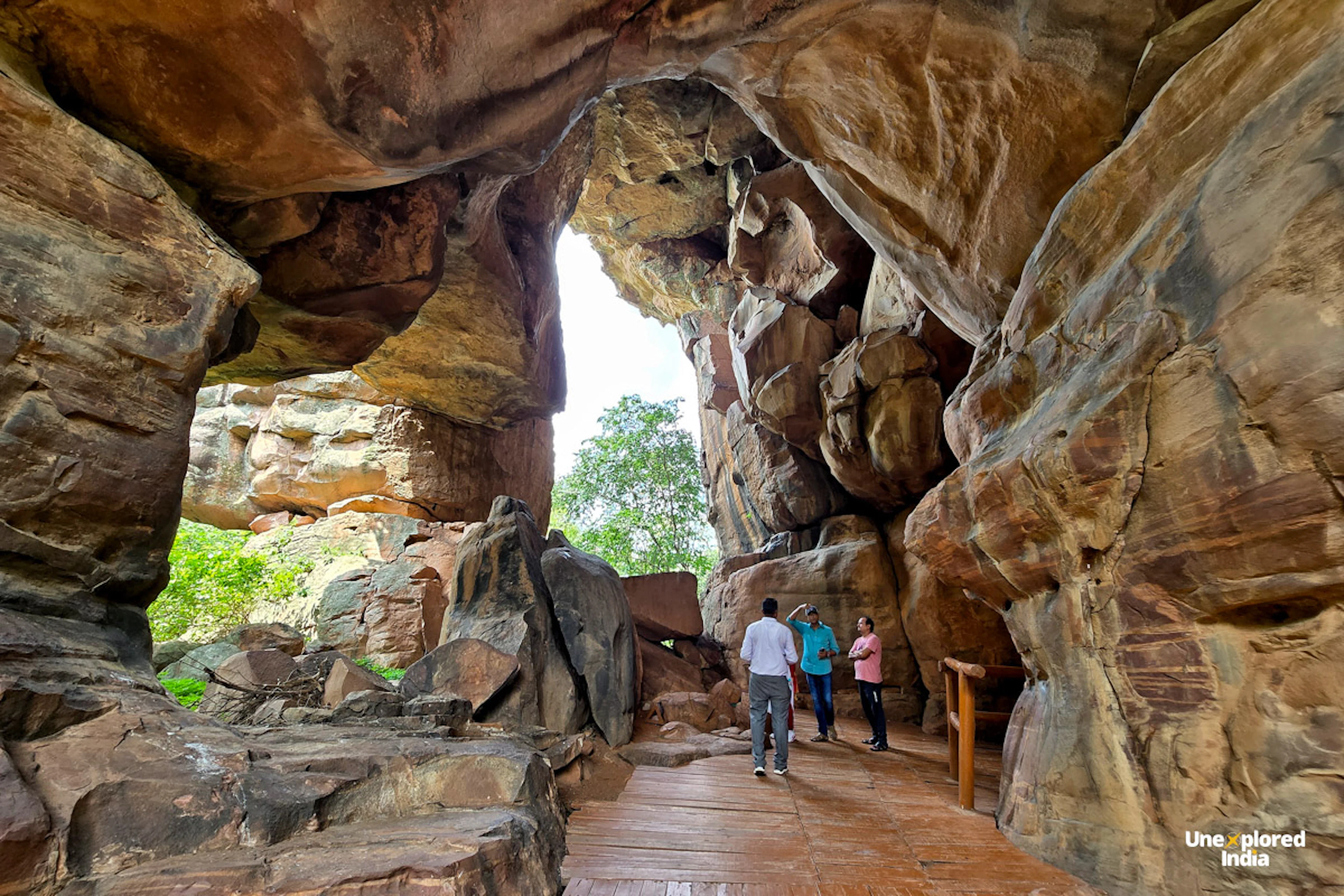 Visitors walk along a paved path through a massive sandstone rock shelter at the Bhimbetka caves, with weathered rock arching overhead and forest visible through the opening.
