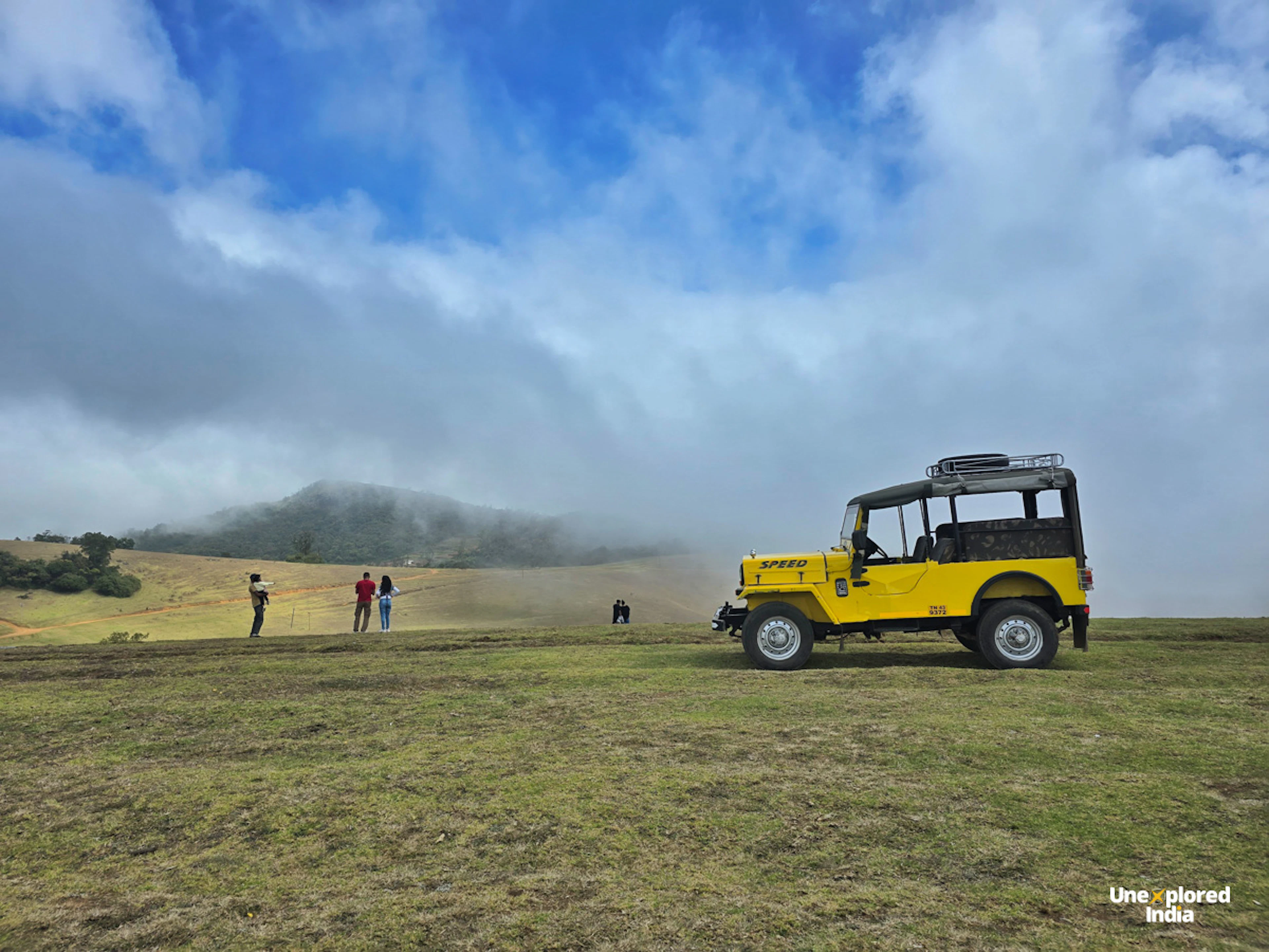 A yellow jeep parked sideways on a vast green grassland. One can see a few people in the background. The sky is covered with clouds.