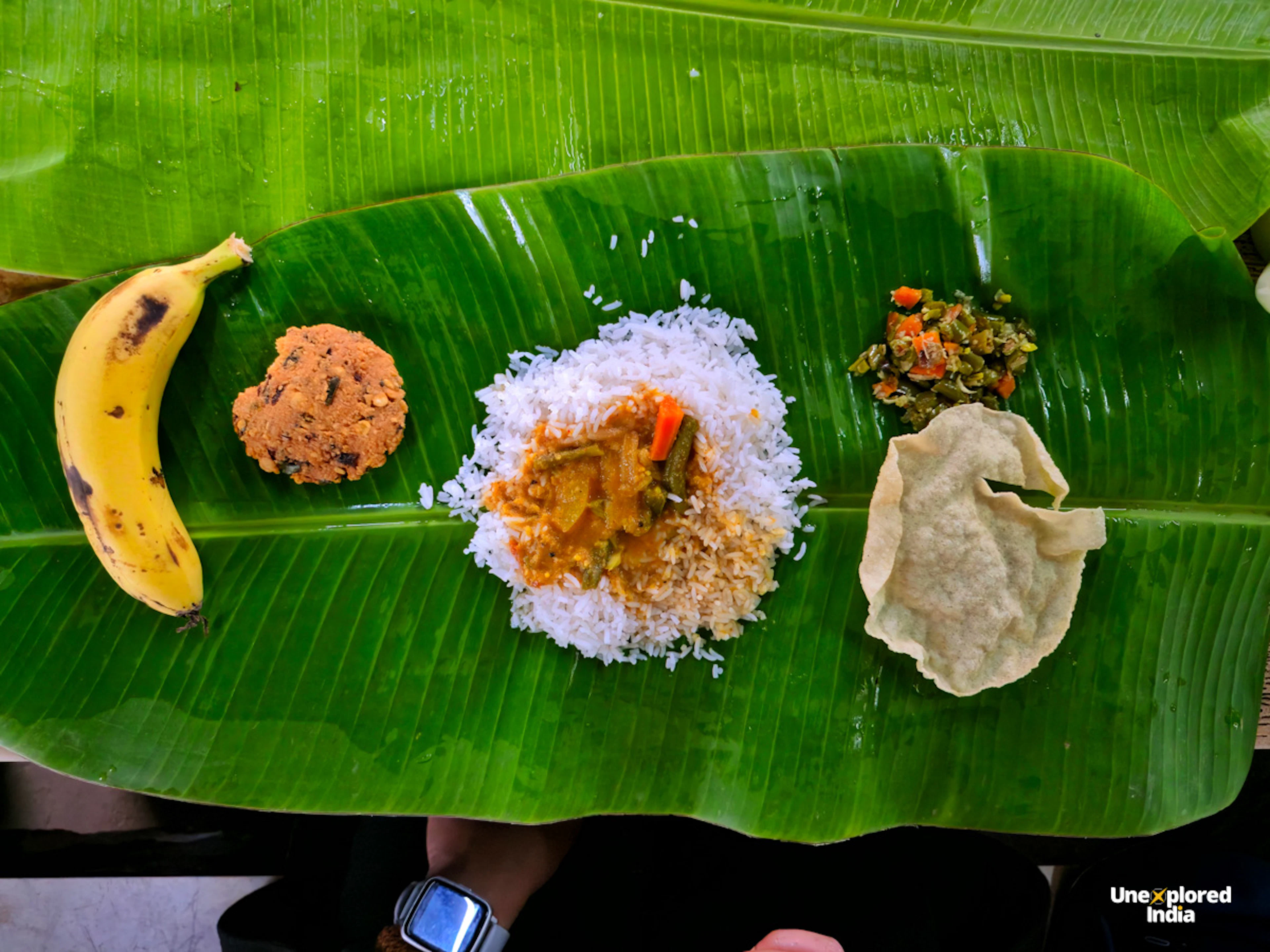 Nilgiris local food served on a banana leaf with rice, curry, vegetables, fritter, papad and banana.