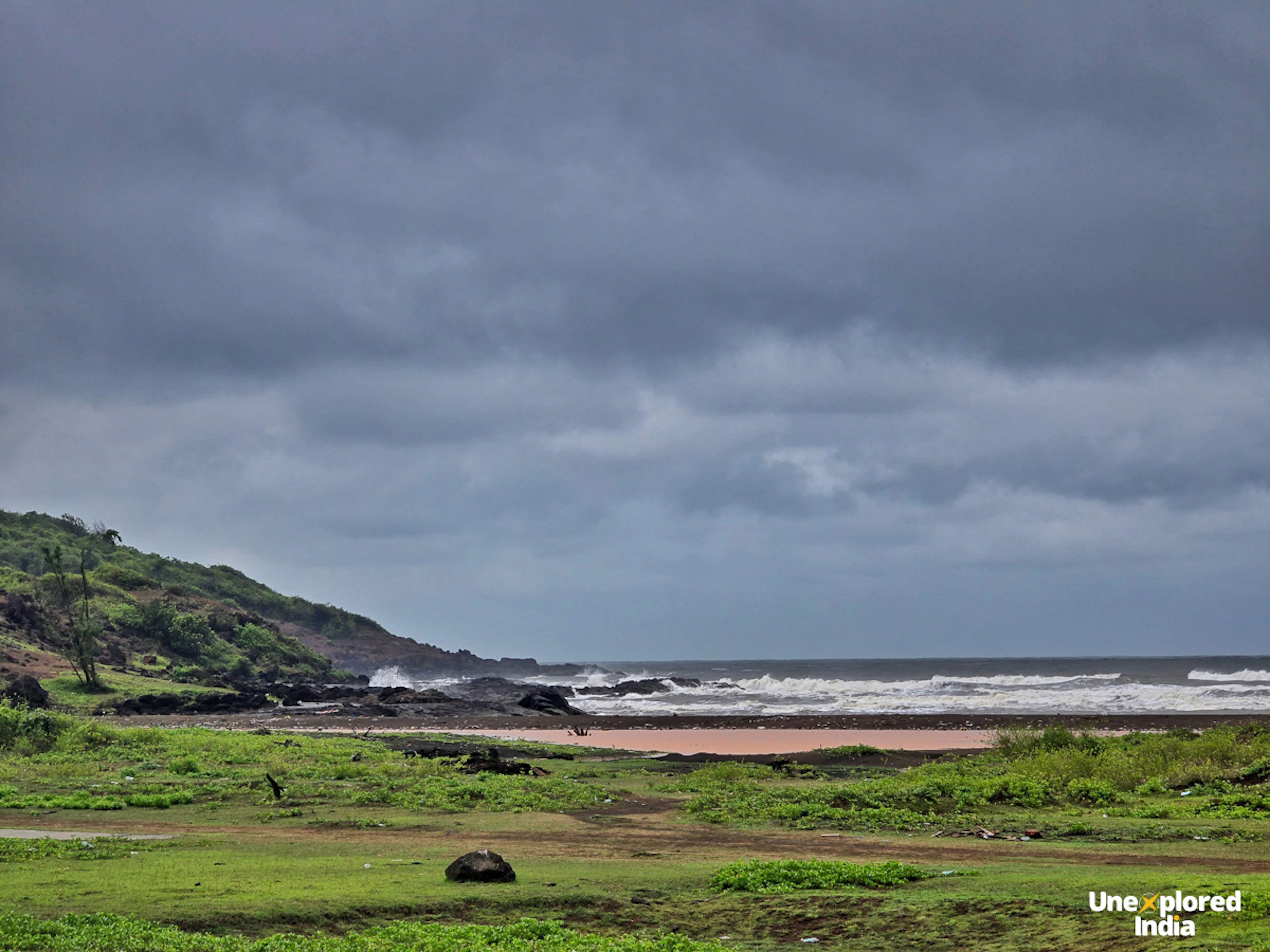 A quiet Ratnagiri beach with waves rolling onto a sandy shore, green hills and coastal vegetation under dark monsoon clouds.