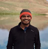 a man in black jacket smiles as he poses for the camera in front of a lake
